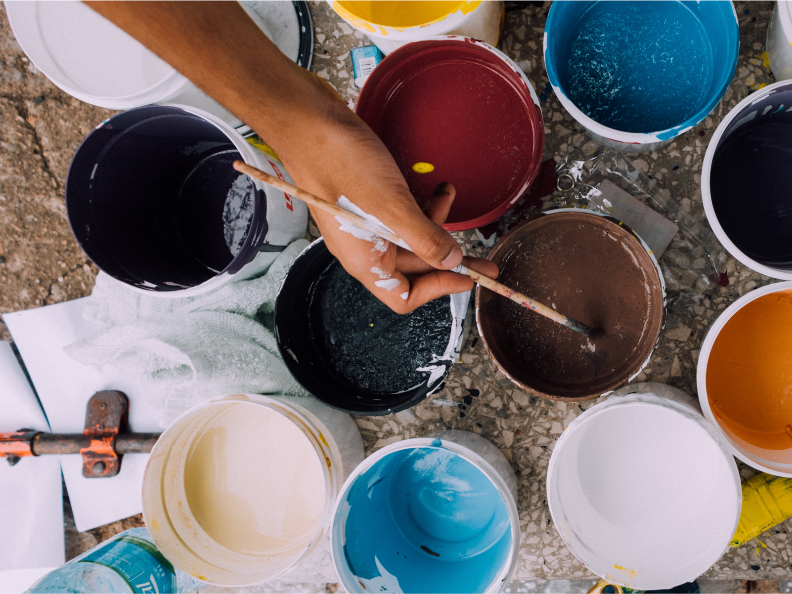 Hand with paintbrush overlooking various paint containers on the floor